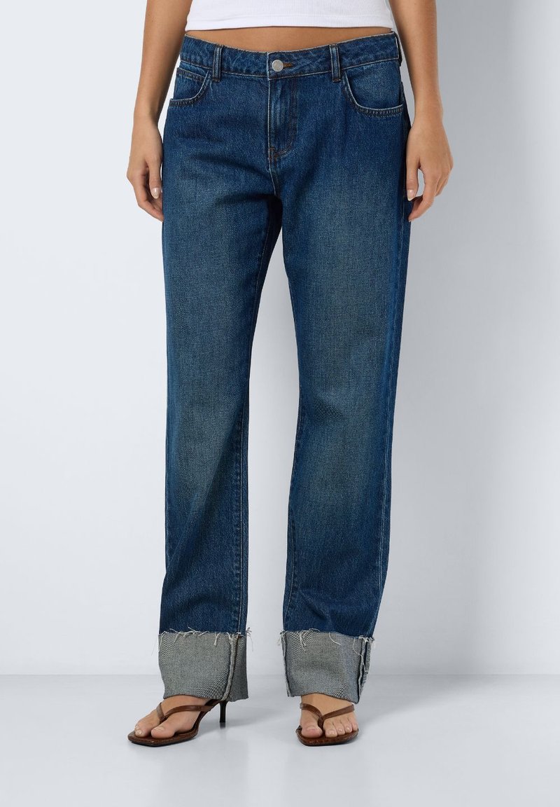 Jeans de ganga em azul escuro com corte reto. Apresenta bainhas dobradas com bordas desfiadas, cinco bolsos e um fecho clássico de botão.