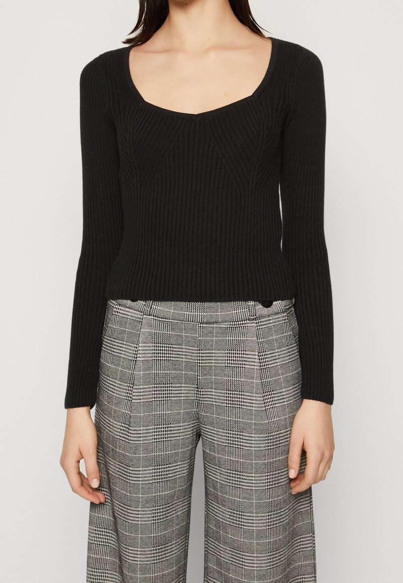 Czarna dopasowana bluzka z długim rękawem i głębokim dekoltem w serek, zestawiona z szarymi spodniami w kratę o wysokim stanie, z szerokimi nogawkami i plisami z przodu.