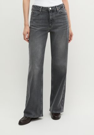 Person trägt hochtaillierte, weit geschnittene, ausgewaschene schwarze Jeans mit Vordertaschen, Knopf und Reißverschluss, kombiniert mit dunklen Schuhen und weißem Oberteil.