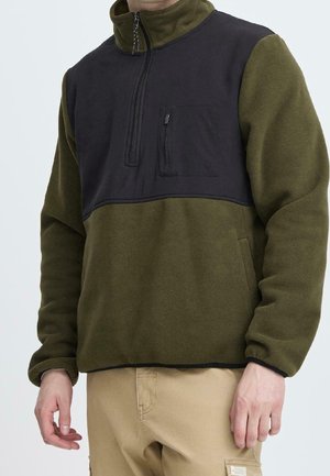 Homme portant une veste polaire vert olive avec un panneau noir sur la poitrine, une poche zippée noire et un pantalon beige.