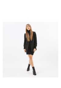 Blazer nero con silhouette aderente, abbinato a un dolcevita nero e a un vestito corto. Indossato con stivaletti neri stringati alla caviglia. Texture liscia.