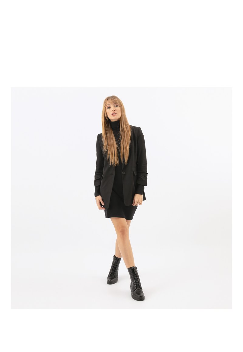 Blazer nero con silhouette aderente, abbinato a un dolcevita nero e a un vestito corto. Indossato con stivaletti neri stringati alla caviglia. Texture liscia.