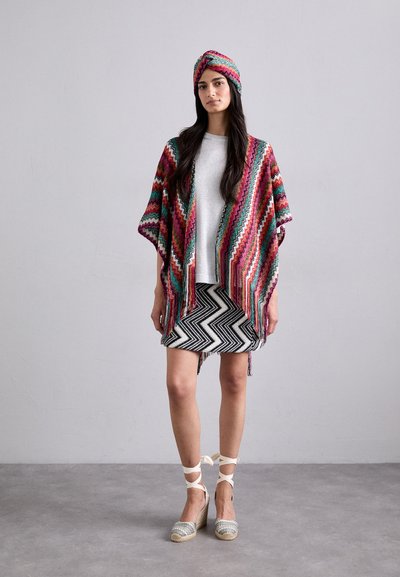 Kimono multicolor, cu franjuri și un model în zig-zag, asociat cu un top argintiu și o fustă în dungi alb-negru. Pantofi espadrile cu curele.