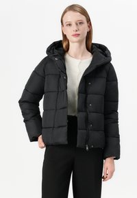 Casaco puffer preto com capuz, design acolchoado e fecho de correr na frente. Textura suave e ajuste casual. Combinado com um top de malha leve e calças escuras.