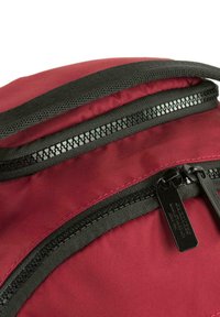 Mochila de tela burdeos con una superficie texturizada, que presenta dobles cremalleras negras y tiradores. Se aprecian bordes cosidos y detalles en contraste.