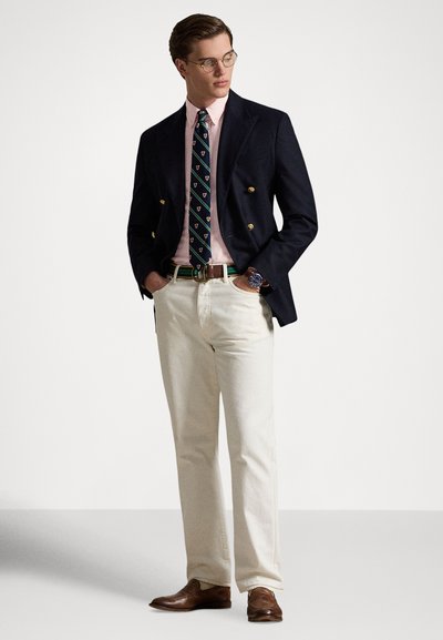 Σκούρο μπλε σακάκι διπλοκουμπωτό, ροζ πουκάμισο με ρίγες, γραβάτα με σχέδια, εκρού παντελόνι, καφέ δερμάτινα παπούτσια, ζώνη με πράσινη λεπτομέρεια, στρογγυλά γυαλιά.