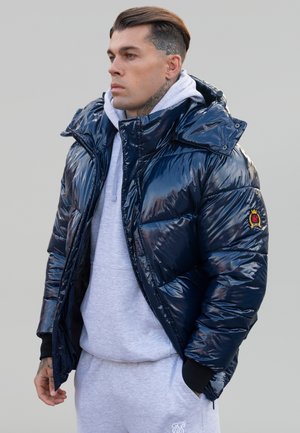 Granatowa kurtka puchowa o błyszczącej powierzchni, z wysokim kołnierzem i kapturem, noszona na jasnoszarym hoodie i dresach. Akcentowana logo marki.