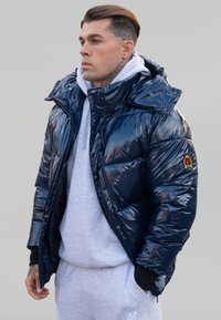 Navy blue puffer jacket with shiny finish and a hood, featuring a zip closure and logo patch, worn over a light gray hoodie and sweatpants.