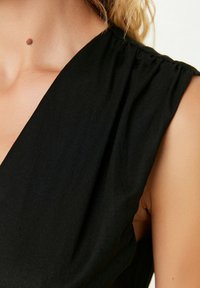 Top noir sans manches avec un décolleté en V profond et des détails plissés aux épaules, présentant une texture de tissu lisse et mate.