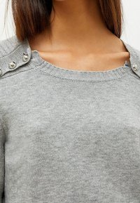 Pull en maille grise avec un col rond, des poignets côtelés et des boutons argentés décoratifs à l'épaule. Texture douce avec un design décontracté.