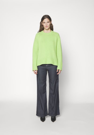 Un pull tricoté vert vif avec un col rond, des poignets côtelés et une coupe ample, associé à un jean noir évasé et des chaussures noires à bouts pointus.