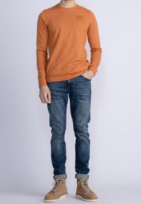 Långärmad orange T-shirt med rund halsringning och en liten logopatch, kombinerad med blå jeans och beige stövlar.