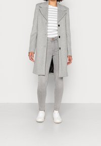 Lichtgrijze wollen jas met een begrijpend kraag en drie knopen, gedragen over een gestreept shirt, gecombineerd met lichtgrijze jeans en witte sneakers.