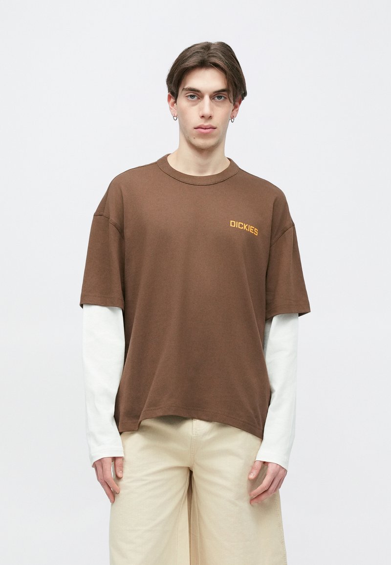 Jeune homme portant un t-shirt Dickies marron superposé à une chemise blanche à manches longues et un pantalon beige clair, debout devant un fond blanc.