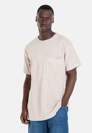 Light beige short-sleeve t-shirt in soft texture with a front pocket and a small logo label at the hem, paired with blue denim shorts.