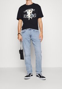 Camiseta negra de manga corta con estampado gráfico; jeans rectos de denim azul claro; zapatillas negras; sosteniendo un pequeño bolso negro.