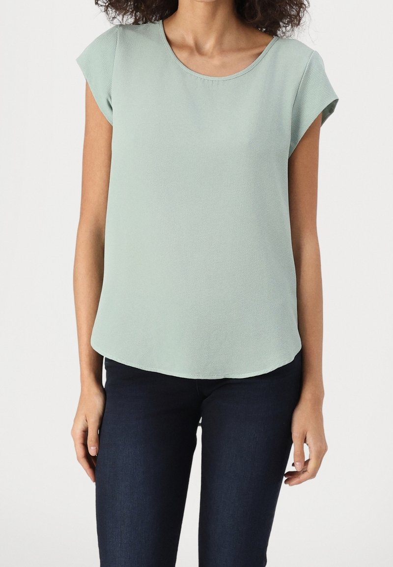 Femme portant un chemisier vert clair à manches courtes et un pantalon bleu foncé ajusté, debout devant un fond uni.