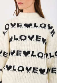 Pletený sveter v krémovej farbe s čiernym textom "LOVE" a vzormi srdiečok. Dizajn s vysokým golierom a textúrovaným povrchom, ktorý má voľný strih.