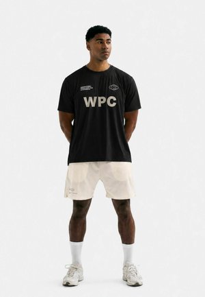 Hombre de pie con las manos detrás de la espalda, vistiendo una camiseta negra WPC, pantalones cortos blancos, calcetines blancos y zapatillas blancas sobre un fondo liso.