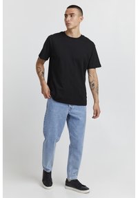 Black cotton t-shirt with short sleeves, paired with light blue denim jeans. Model wears black sneakers and showcases visible tattoos on arms.