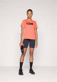 Neono oranžinė sportinė marškinė su juodu užrašu "LDN", derinami su tamsiomis šortais ir juodais bėgimo batais, laikant juodą vandens butelį.