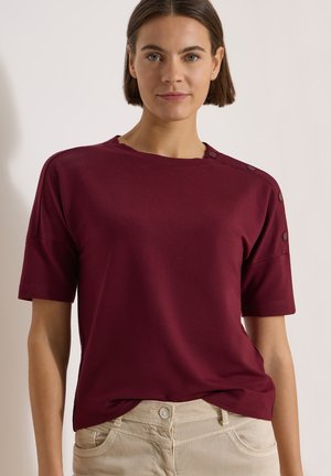 Femme aux cheveux courts et bruns portant un haut bordeaux à manches courtes avec des boutons sur l'épaule et un pantalon beige, debout devant un fond neutre.
