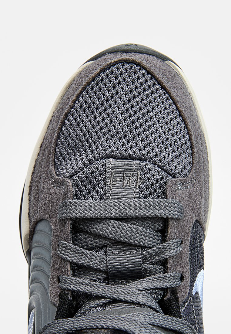 Primer plano del antepié de una zapatilla deportiva gris con parte superior de malla, acentos de ante y cordones gris oscuro bien atados, sobre fondo blanco.