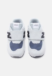 Γκρι σουέτ αθλητικά παπούτσια με σκούρες μπλε сетρί λωρίδες, διαθέτουν λουράκι με βέλκρο και το χαρακτηριστικό λογότυπο «N» στο πλάι. Κλασικός σχεδιασμός αθλητικού παπουτσιού.