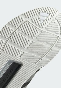 Semelle de chaussure de sport grise et blanche avec des rainures et motifs texturés, présentant un panneau noir avec le texte "REPETITOR" et un logo triangulaire.