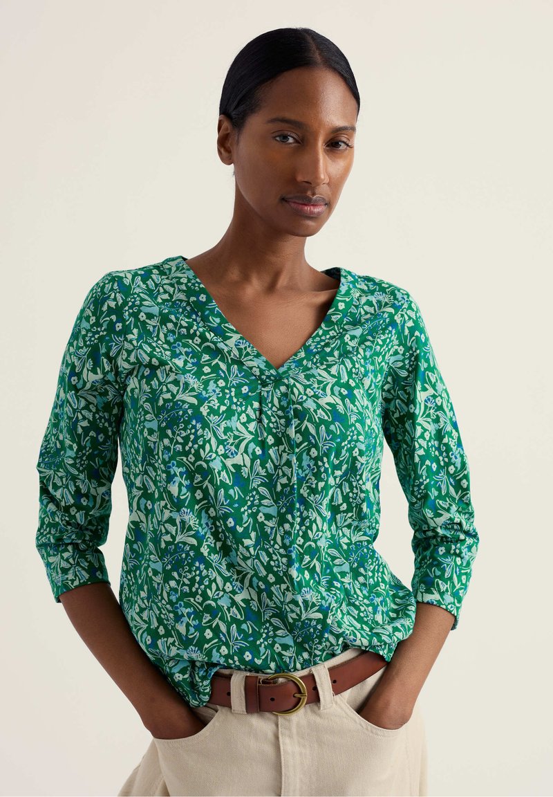 Frau mit dunklen Haaren in einer grünen Bluse mit V-Ausschnitt und Blumenmuster sowie beigen Hosen, stehend mit den Händen in den Taschen vor einfarbigem Hintergrund.