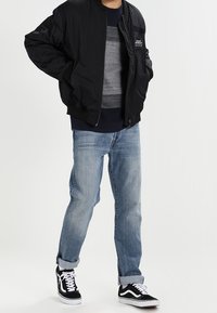 En person som bär en svart bomberjacka, grå randig tröja, ljusblå jeans med uppvikta ben och svart-vita sneakers står med händerna i fickorna.