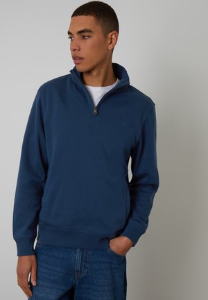 Junger Mann trägt einen marineblauen Quarter-Zip-Sweatshirt über einem weißen Hemd und blauen Jeans, schaut nach links mit einer Hand in der Tasche.