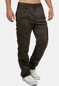 Olivgrüne Cargo-Hosen mit mehreren Reißverschluss- und Klappentaschen, einer strukturierten Oberfläche und einem geraden Schnitt. Getragen mit weißen Sneakers.