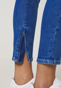Jeans de denim azul con cremalleras en los tobillos, que cuentan con un ajuste slim y una longitud recortada. El tejido tiene una textura suave y un desgaste sutil.