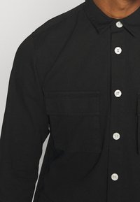 Zwarte overhemd met knopen en kraag, voorzien van twee borstzakken en witte knopen, gemaakt van een gladde stof, met een losse pasvorm.