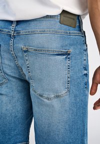 Jasnoniebieskie dżinsy z klasycznym pięciopocketowym designem, widocznym przeszyciem oraz skórzaną łatą na pasku.