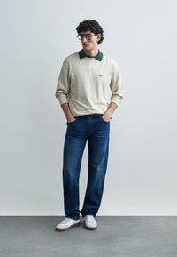 Beige long-sleeve polo with a dark green collar, worn with blue jeans and white sneakers. Model has curly hair and glasses.