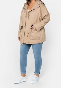 Beige leichte Jacke mit Kapuze, ausgestattet mit Kordeln in der Taille, einem Frontreißverschluss, Druckknöpfen und zwei Vordertaschen. Getragen über einem beigen Oberteil.