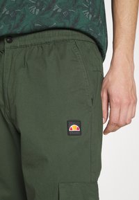 Un primer plano de una mano de una persona descansando sobre pantalones verde oliva que presentan un parche de marca. La camisa tiene un patrón floral en tonos verdes.
