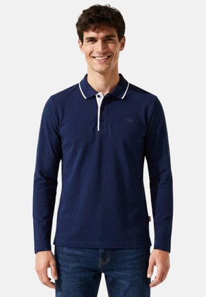 Jeune homme souriant portant un polo à manches longues bleu marine avec un bord blanc sur le col et un jean bleu foncé.