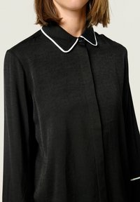 Black blouse featuring a textured fabric, white piped collar, and button closure. Long sleeves with a relaxed fit for ease of movement.