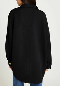 Svart oversized skjorta, texturerat tyg med diamantmönster, långa ärmar, knäppta manschetter, böjd fåll, subtil detalj med gyllene knappar.