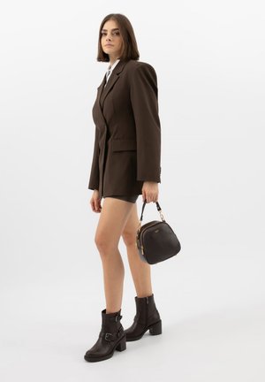 Mladá žena v tmavě hnědých šatech s blazerem a kotníkovými botami drží černou koženou kabelku na bílém pozadí.
