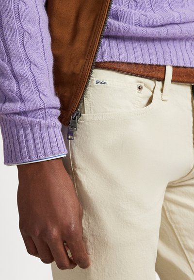 Ljubičasti džemper s pletenim kabel uzorkom ispod smeđe jakne od brušene kože, u kombinaciji sa svijetlo bež hlačama. Ima patentni zatvarač i oznaku "Polo" na džepu.