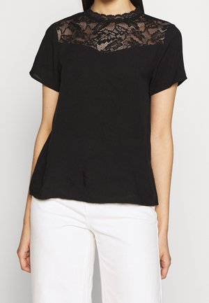 Vrouw draagt een zwarte blouse met korte mouwen en kanten details op de bovenborst en schouders, gecombineerd met een witte broek.