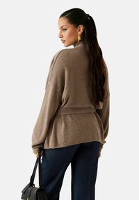 Maglione marrone lavorato a maglia con cintura in vita, spalle scese e vestibilità comoda, abbinato a pantaloni blu scuro e una borsa nera.