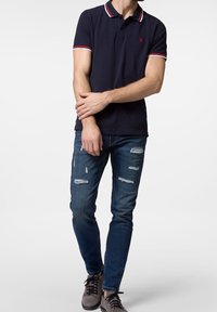 Mörkblå polo med röd och vit randig krage, kombinerad med slitna blå jeans. Modellen bär grå sneakers och har händerna korsade runt midjan.