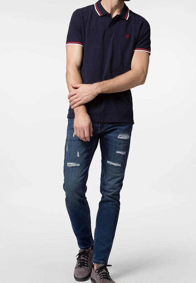 Mörkblå polo med röd och vit randig krage, kombinerad med slitna blå jeans. Modellen bär grå sneakers och har händerna korsade runt midjan.