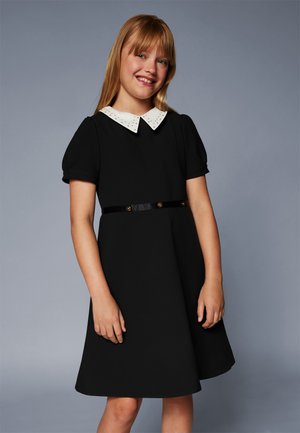 Musta mekko lyhyillä puhvihihnoilla, valkoinen kaulus niitteihin koristeltuna ja musta vyö, jossa on kultaiset napit, esitelty harmaata taustaa vasten.