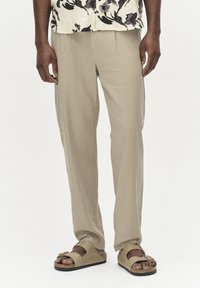 Pantalons en lin beige clair avec une coupe décontract�ée, dotés d'une poche latérale. Associés à des sandales beiges confortables. Chemise à manches courtes à motifs floraux visible.